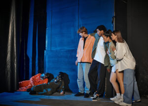 Group of young people watching actors wearing gas masks and costumes, performing in an industrial setting with blue lighting.