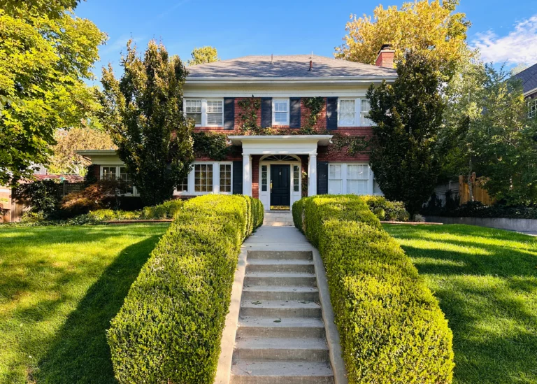 A stately brick Colonial house in Yalecrest, featuring a symmetrical facade with white columns framing the entrance, multi-paned windows with shutters, and a neatly manicured lawn. The home’s traditional design reflects the neighborhood's historic charm, with mature trees lining the street in the background.